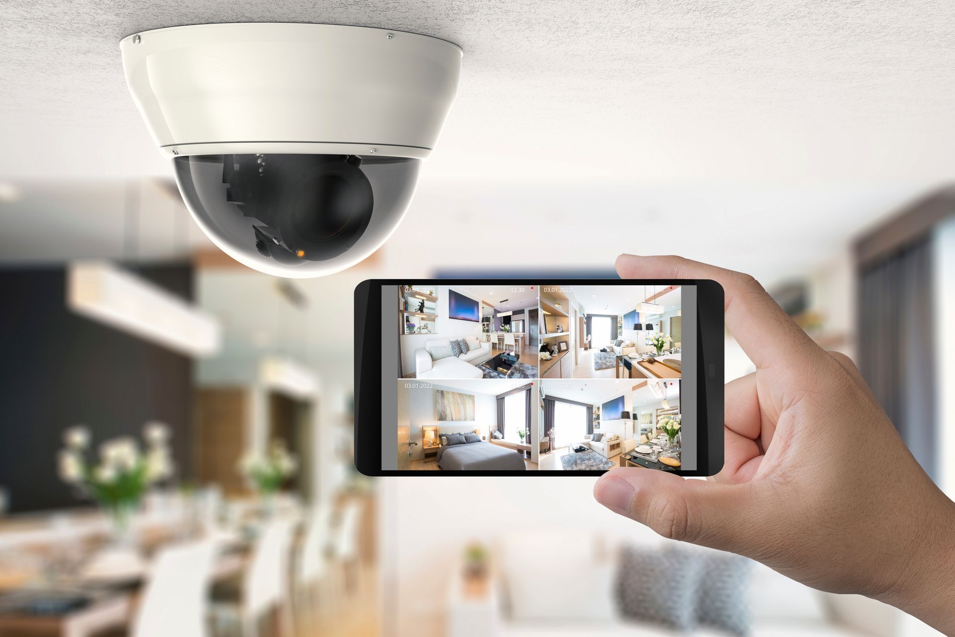 Une main tient un smartphone affichant le flux vidéo multivue d'une caméra de sécurité domestique, la caméra elle-même étant fixée au-dessus.
