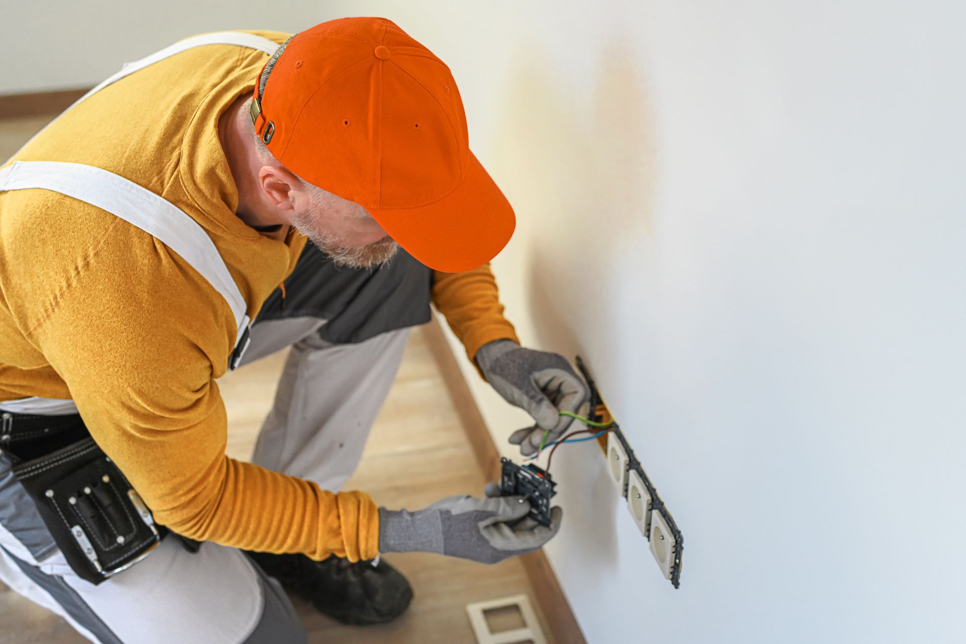 Un électricien en chemise jaune et casquette orange est agenouillé pour installer des câbles électriques dans un panneau mural.
