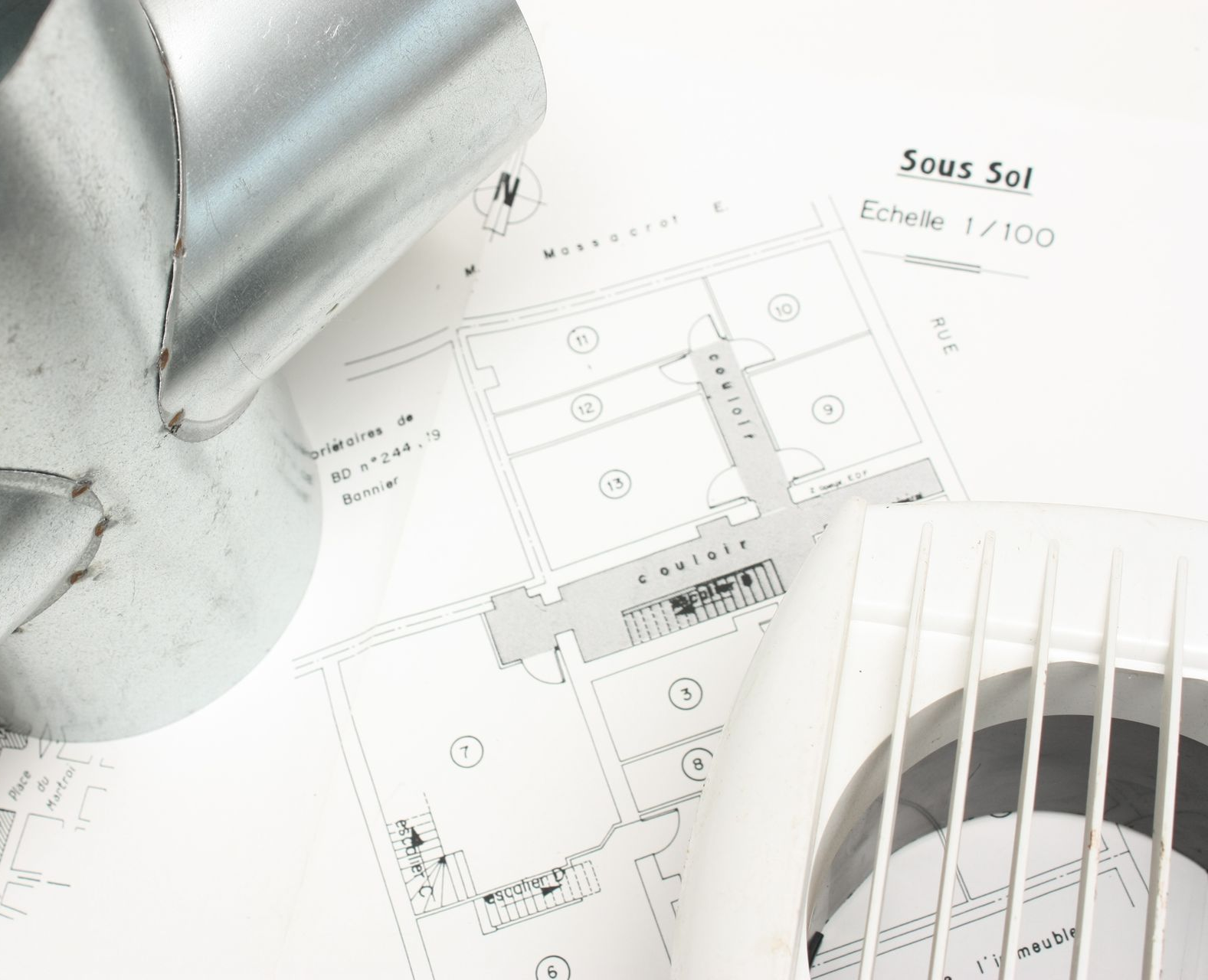 Un plan d'étage de sous-sol repose sur une table, sous un raccord de ventilation métallique et un couvercle de ventilation en plastique blanc.