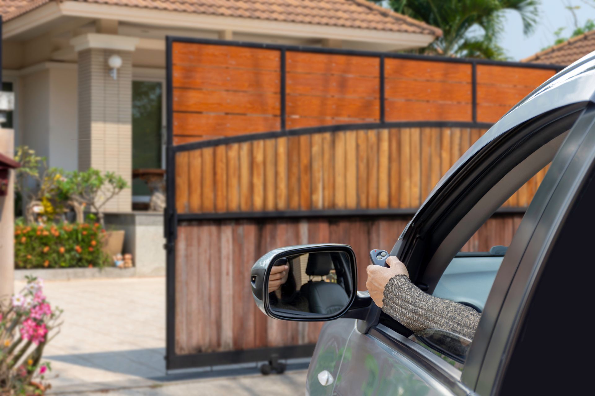 Une personne dans une voiture tient une télécommande pour ouvrir un portail en bois devant une maison.