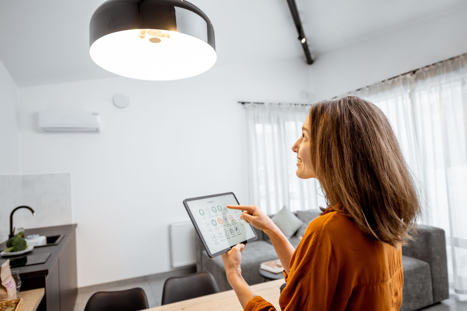 Une personne vêtue d'un haut orange utilise une tablette pour contrôler l'éclairage intelligent d'un salon et d'une cuisine modernes.