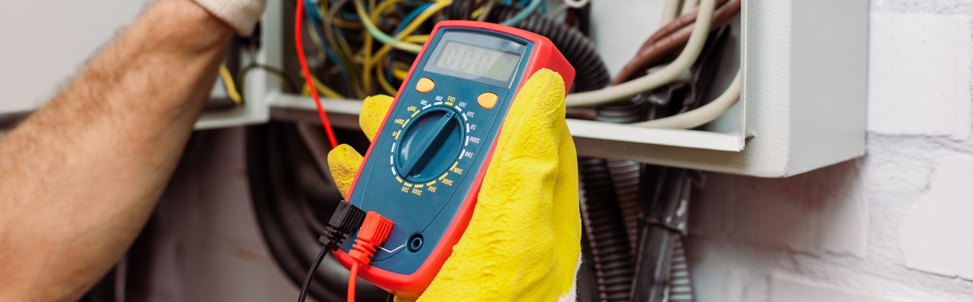 Une main gantée utilise un multimètre numérique pour tester le câblage électrique à l'intérieur d'un boîtier électrique ouvert.