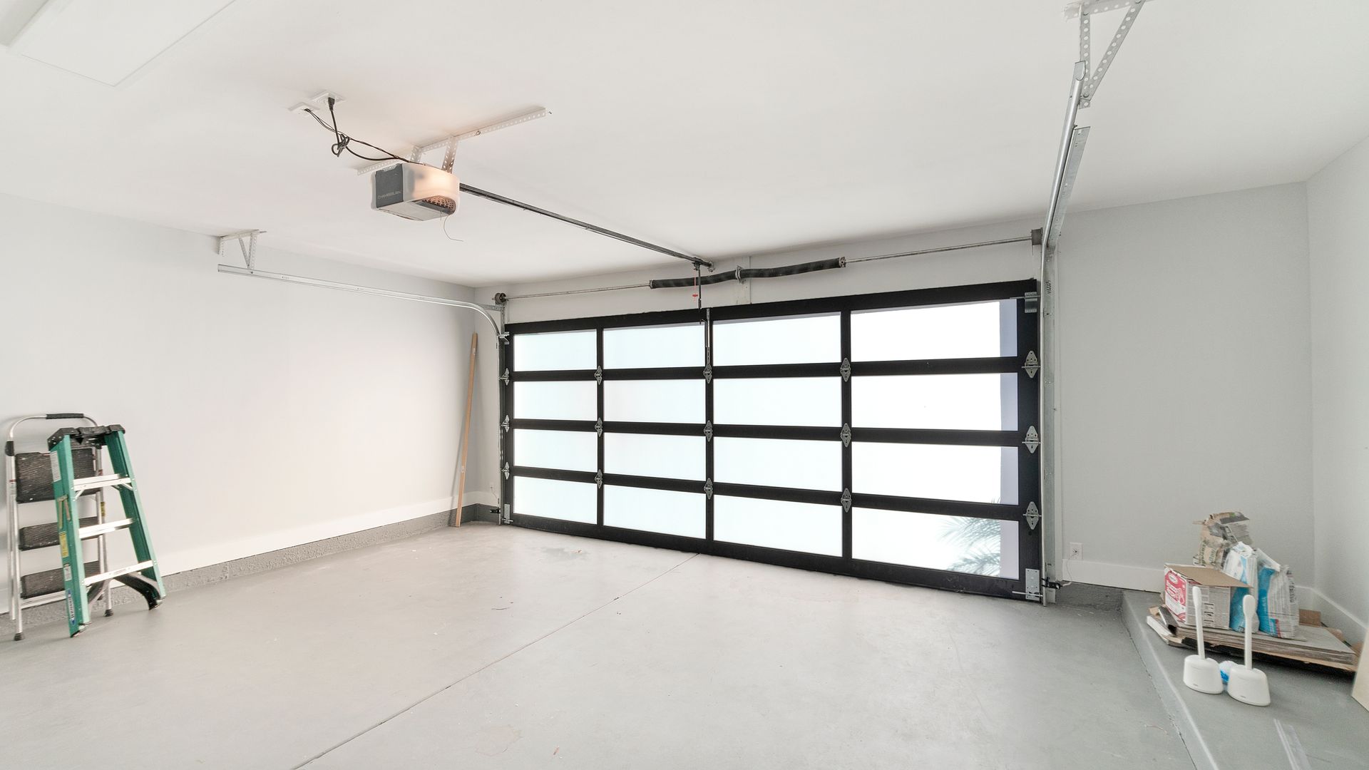 Un garage blanc vide, équipé d'une porte de garage moderne en verre à cadre noir, d'un escabeau et de quelques produits de nettoyage.