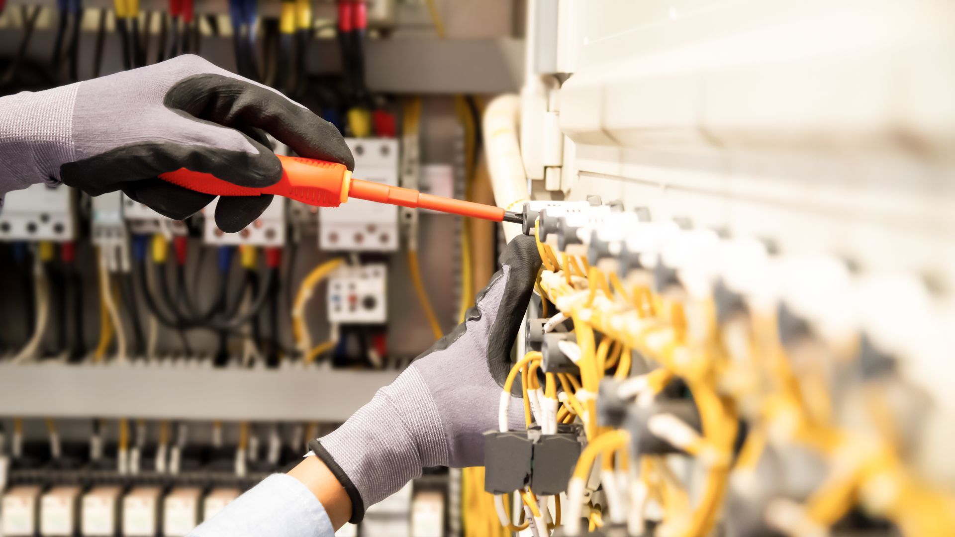 Une personne portant des gants de travail utilise un tournevis à manche orange pour serrer une vis dans un panneau de commande électrique complexe.