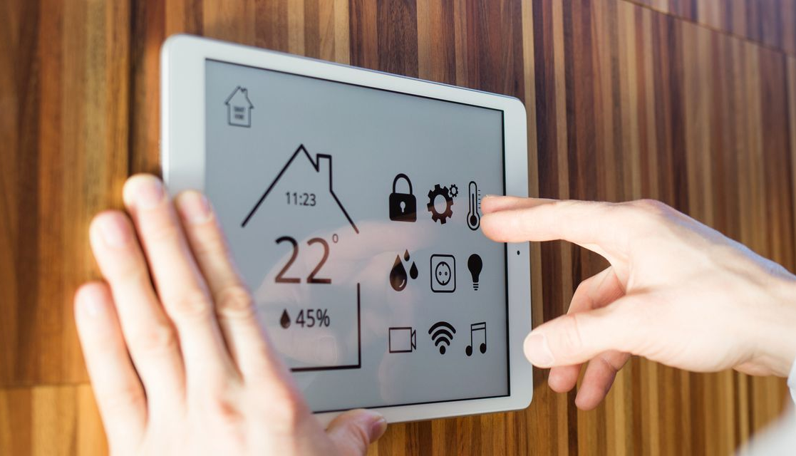 Des mains utilisent une tablette fixée sur un mur en bois pour contrôler un système domotique affichant la température, la sécurité et les paramètres des appareils.