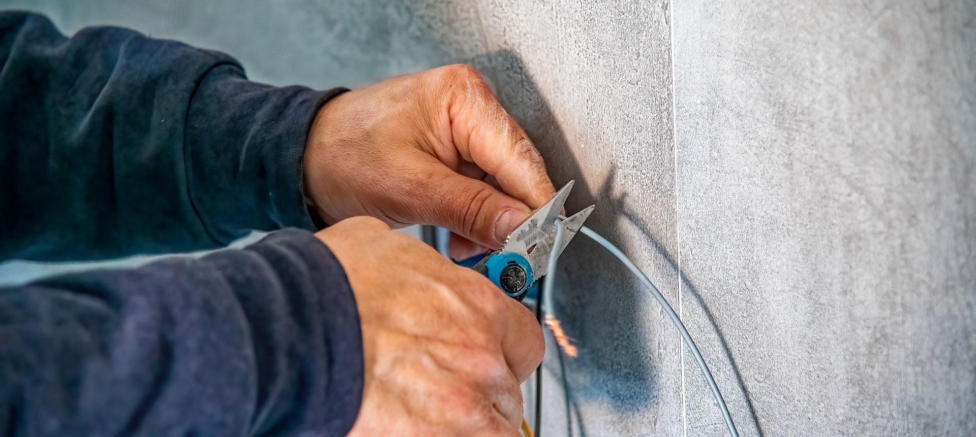 Gros plan sur des mains utilisant une pince coupante pour dénuder l'isolant d'un câble électrique contre un mur en béton gris.