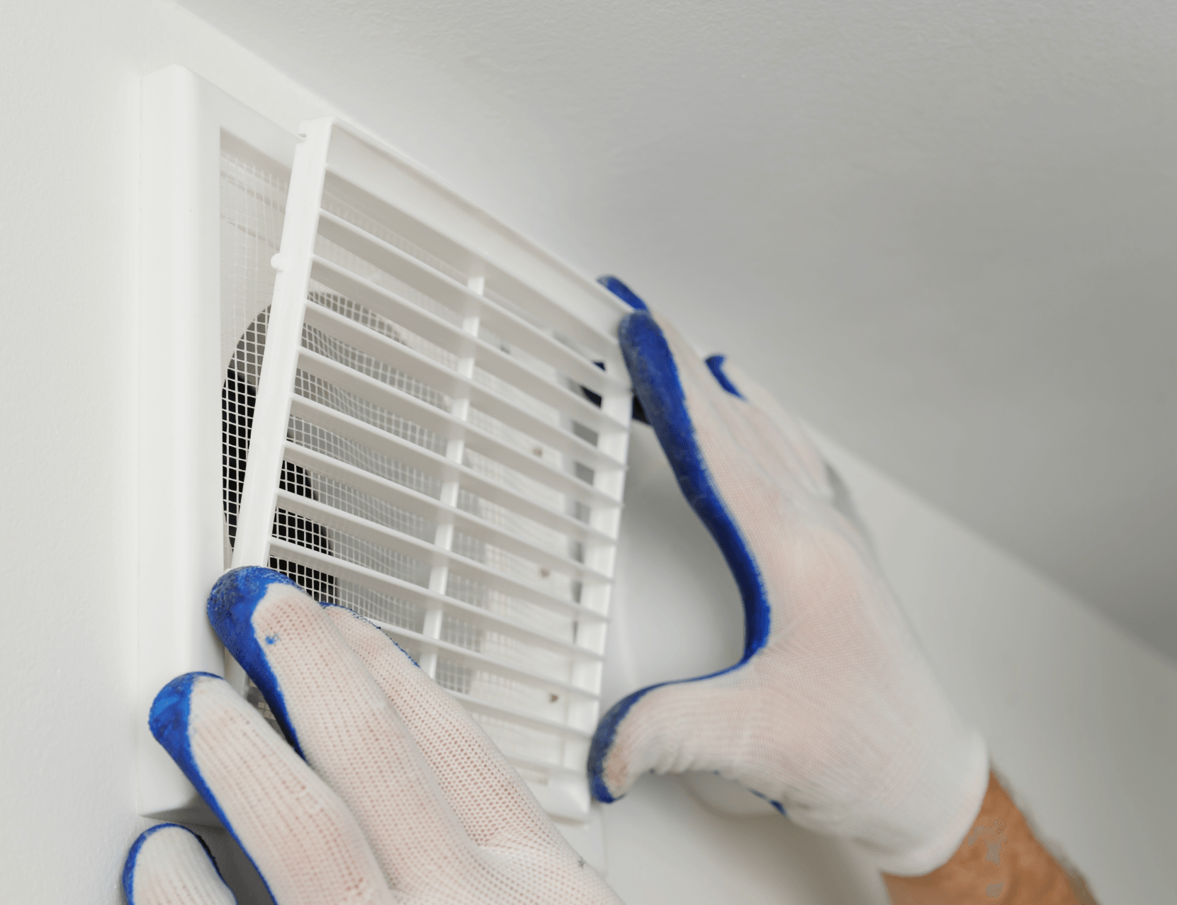 Des mains avec des gants de travail blancs et bleus fixent une grille de ventilation en plastique blanc sur un mur intérieur blanc.