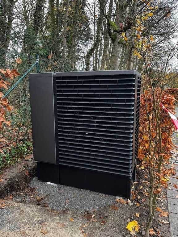Dunkelgraue Wärmepumpeneinheit im Außenbereich, in der Nähe von Bäumen.