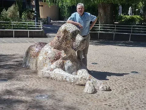 Hombre de pie junto a una gran escultura tipo mosaico de un león reclinado en un parque.