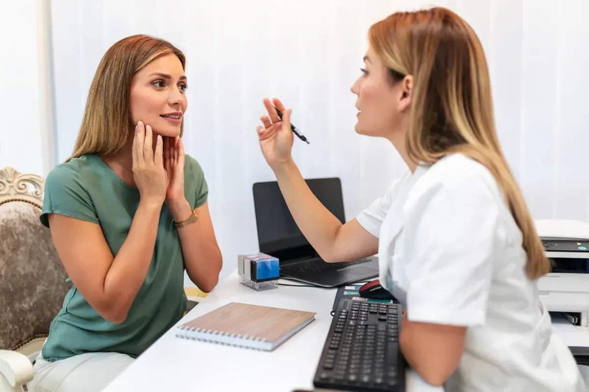 Una mujer consulta con un profesional médico en un escritorio. Uno le toca la garganta, el otro sostiene un bolígrafo