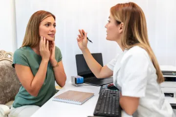 Una mujer consulta con un profesional médico en un escritorio. Uno le toca la garganta, el otro sostiene un bolígrafo