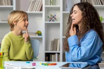 Niño y mujer practicando terapia del habla, ambos señalando sus bocas, en el interior.
