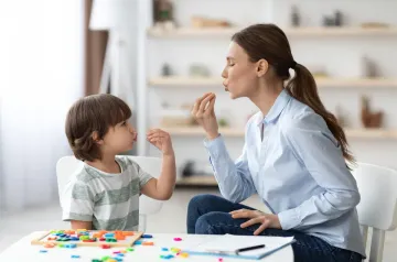 Una mujer enseña a un niño; ambos forman un beso con la boca. Habitación blanca.