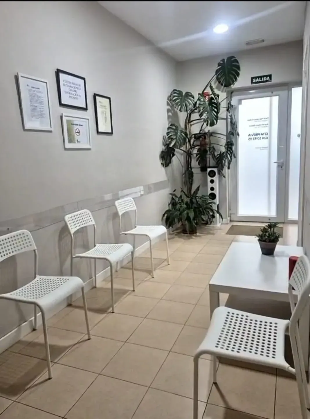 Sala de espera con sillas blancas, mesa, plantas y puerta de salida.