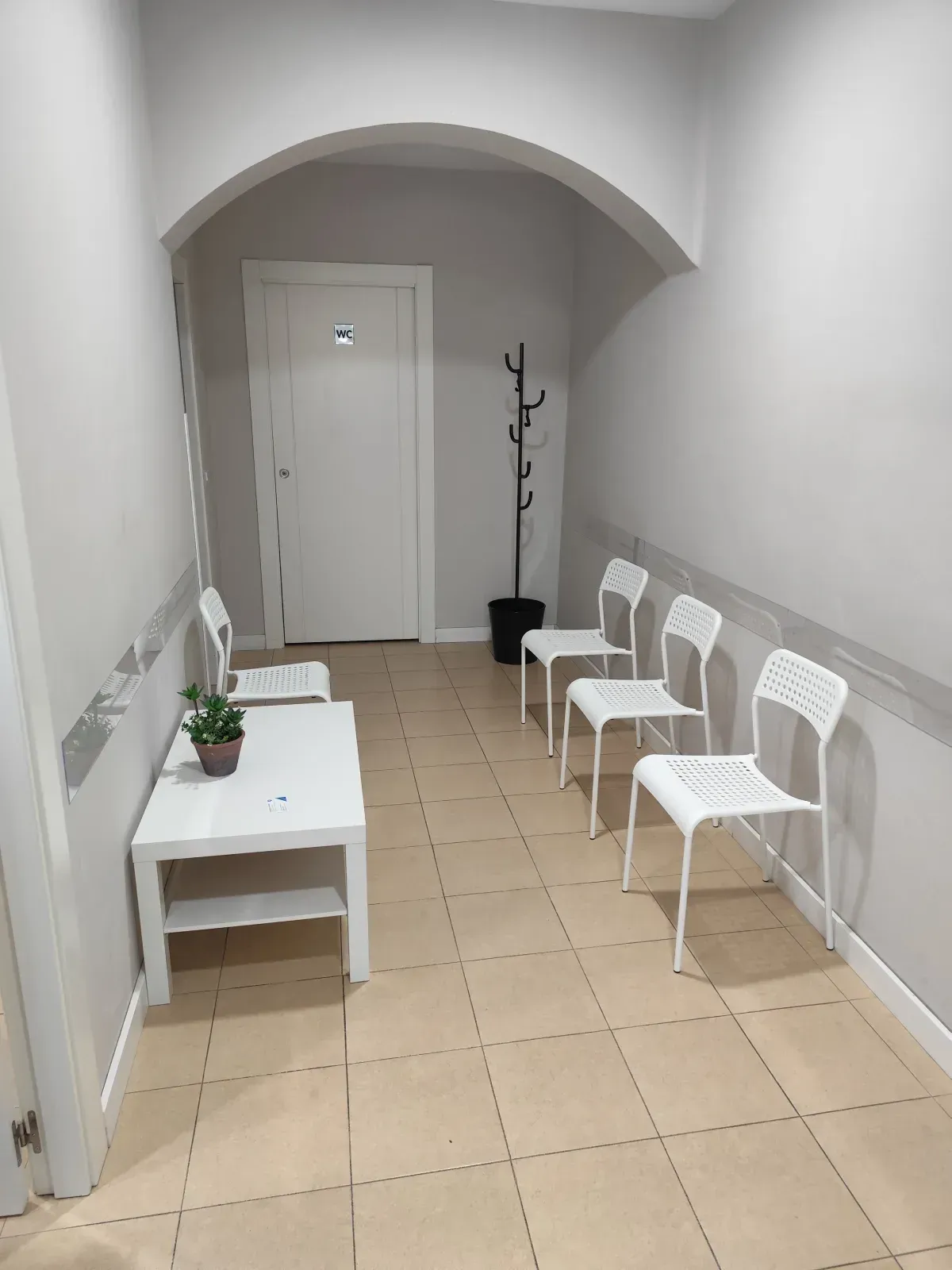 Sala de espera estrecha con sillas, mesa y puerta blancas. Paredes grises y suelo de baldosas.