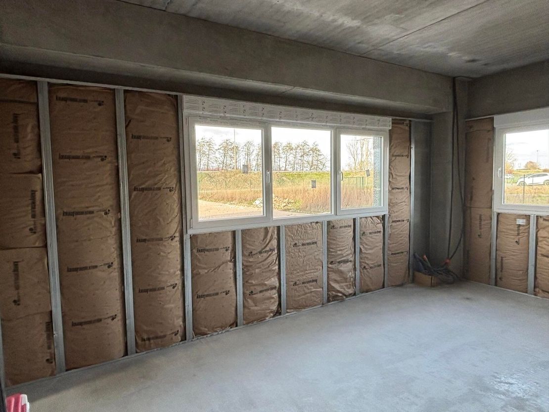 Pièce inachevée avec isolation apparente, des fenêtres et un sol en béton brut.