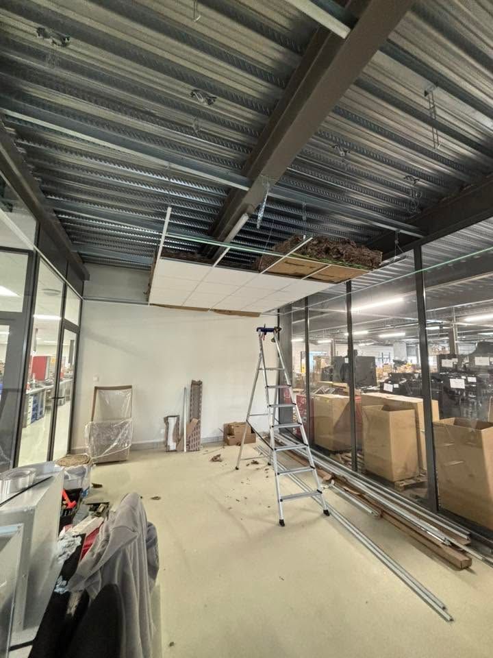 Intérieur d'une façade de magasin en construction avec plafond métallique, des échelles et matériaux empilés derrière des parois vitrées.