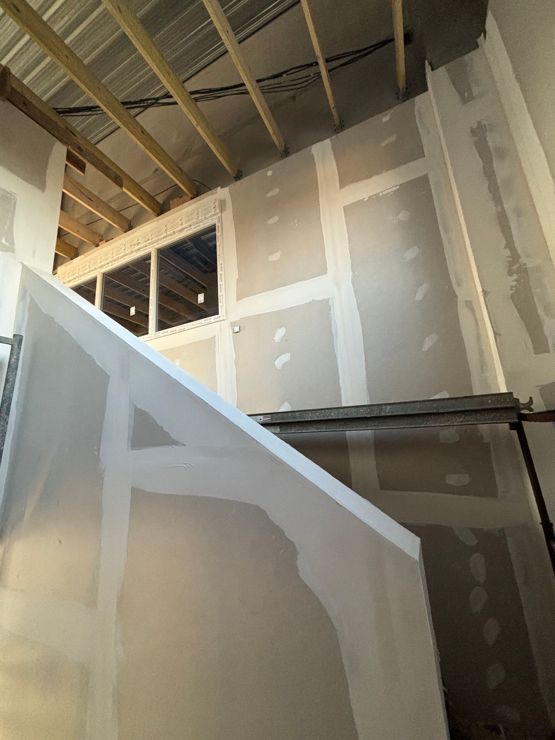 Intérieur d'une cage d'escalier inachevée avec des cloisons sèches, des solives de plafond apparentes et une petite ouverture de fenêtre.