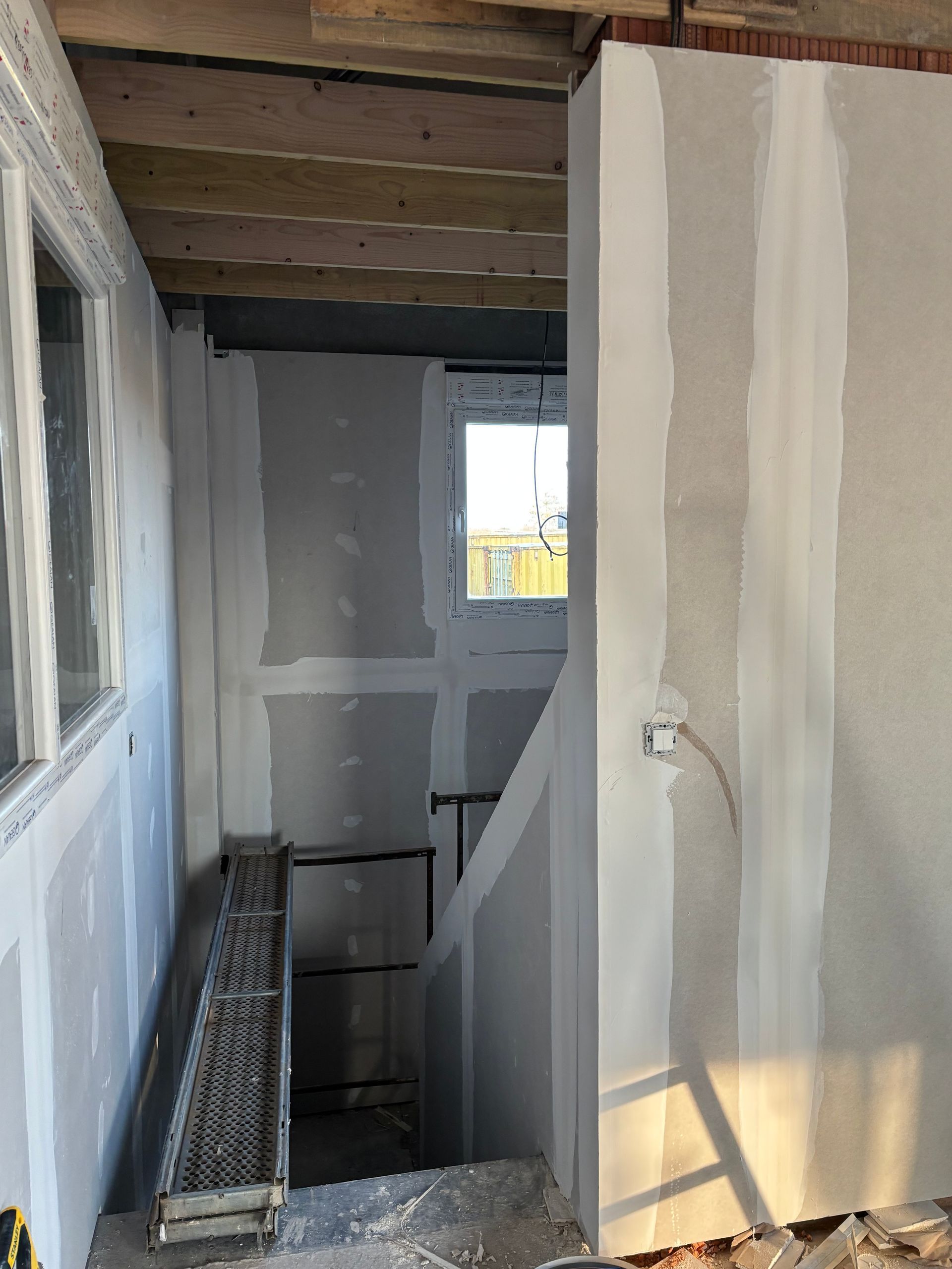 Escalier étroit et inachevé avec  des cloisons sèches, des solives apparentes et une petite fenêtre en bas.