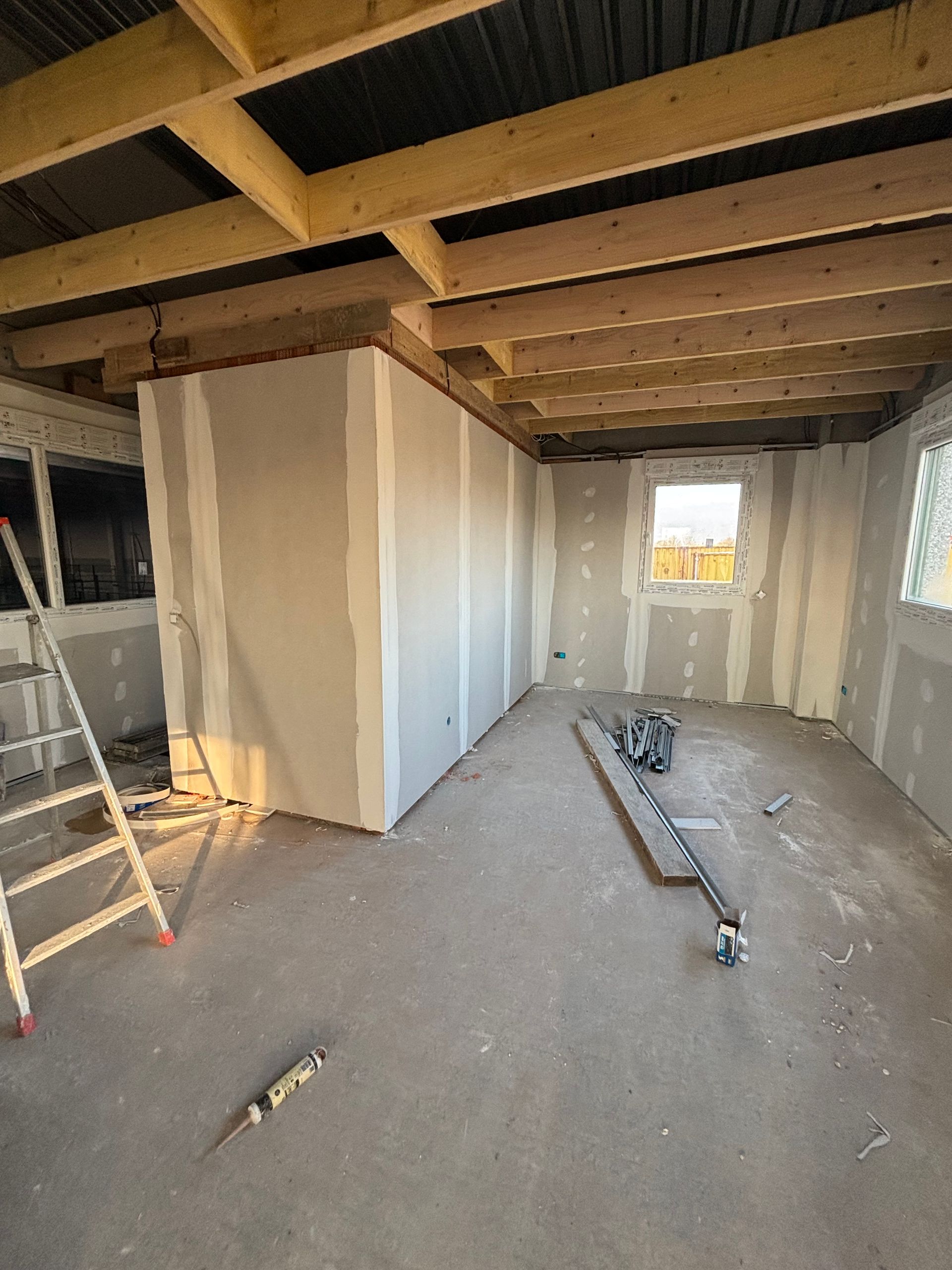 Pièce intérieure inachevée avec poutres apparentes au plafond, murs en plaques de plâtre, échelle et outils de construction au sol.