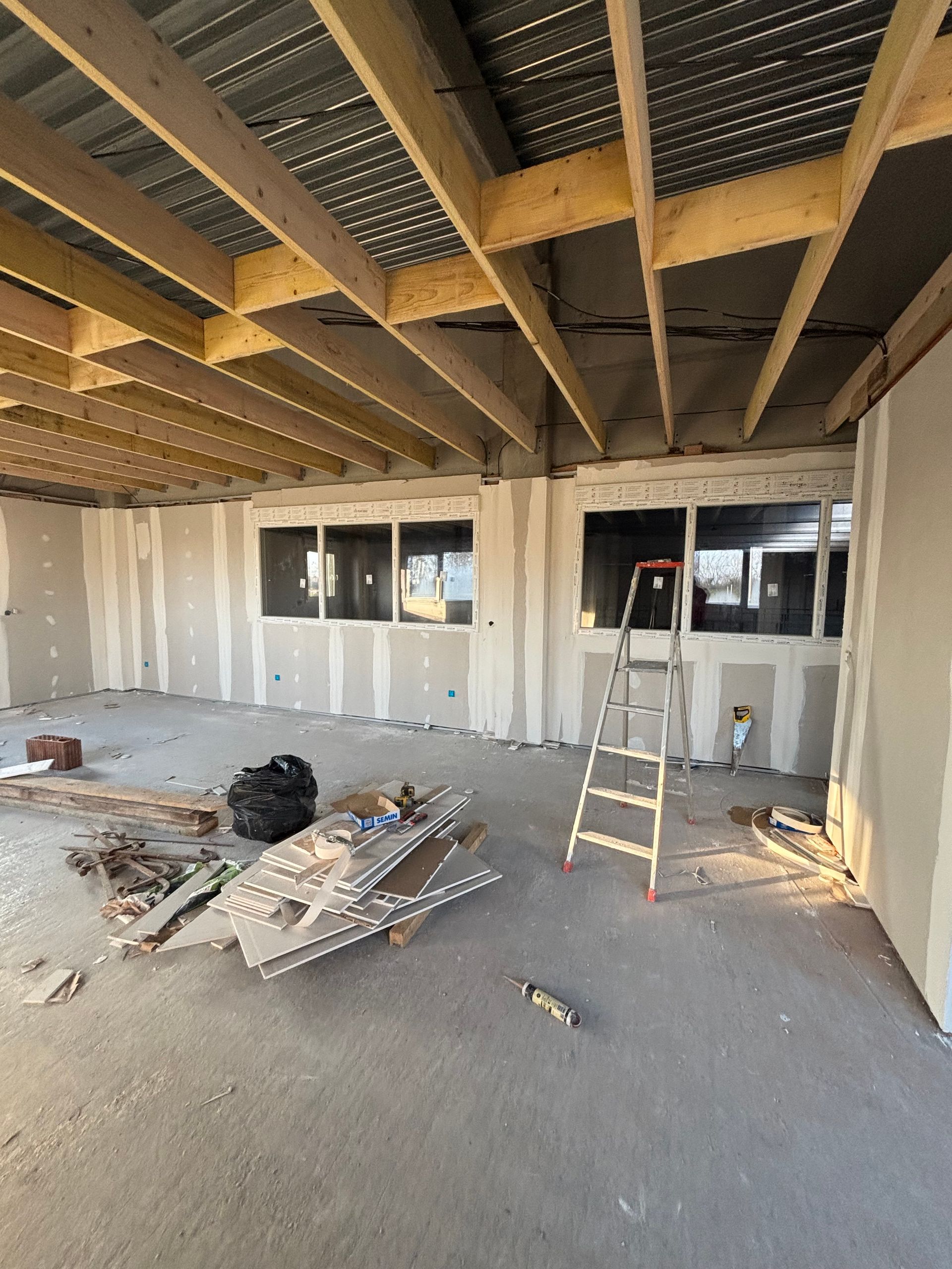 Pièce en rénovation avec plafond apparent, panneaux de plaques de plâtre, tas de débris et une échelle près des fenêtres.