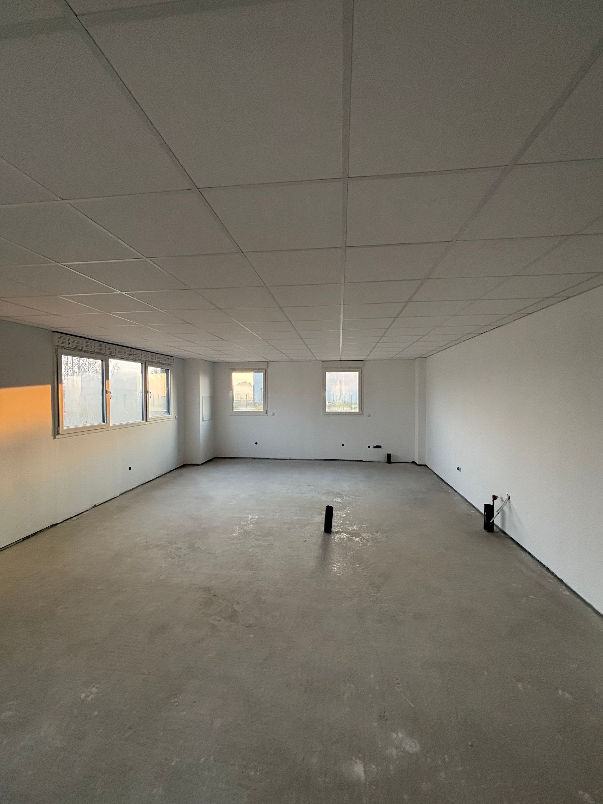 Pièce vide et inachevée avec un sol en béton gris, des murs blancs, un faux plafond et plusieurs fenêtres laissant entrer la lumière.
