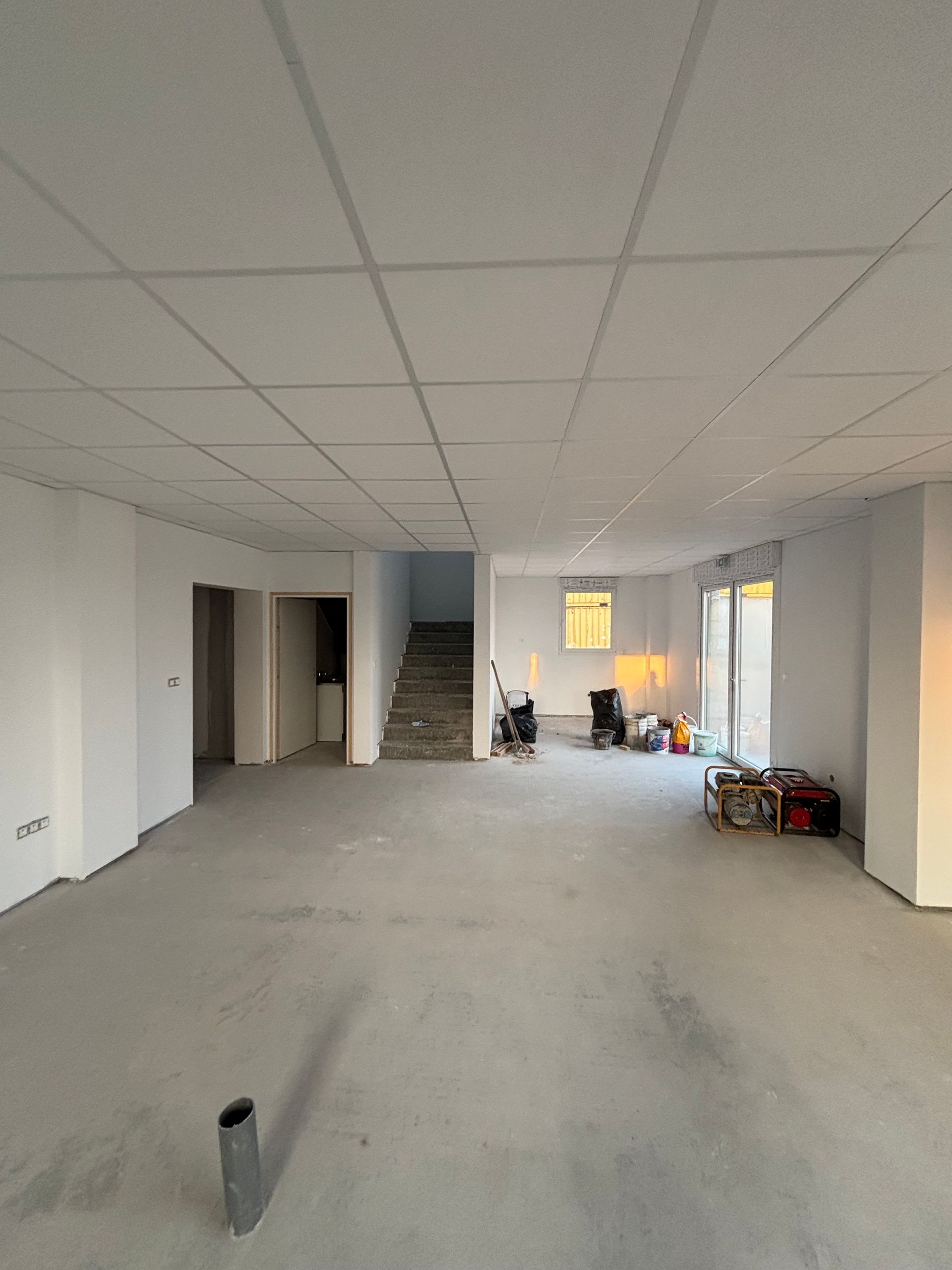 Intérieur vide et inachevé, avec des murs blancs, un sol gris, un faux plafond et des outils de travail près de fenêtres lumineuses.