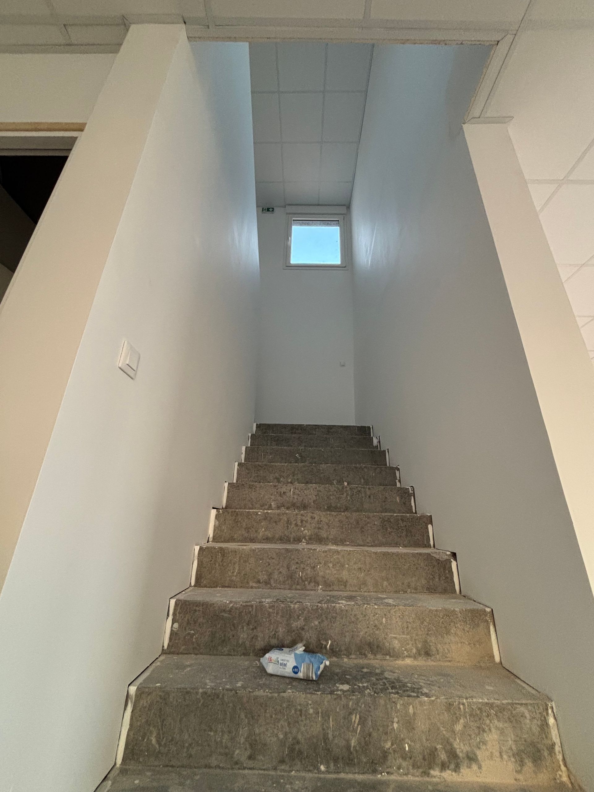 Escalier intérieur étroit aux marches usées, des murs blancs et une petite fenêtre en haut.