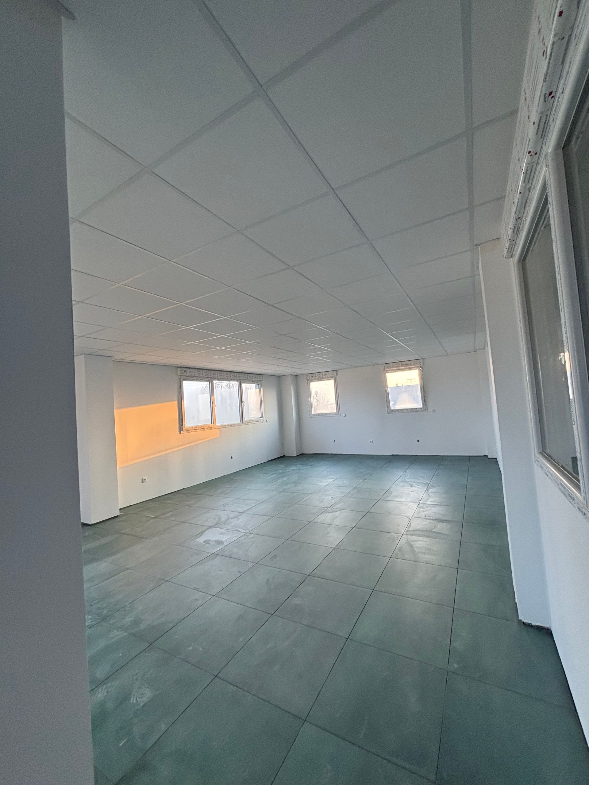 Chambre vide non meublée avec de grandes fenêtres, des murs blancs et un sol carrelé vert.