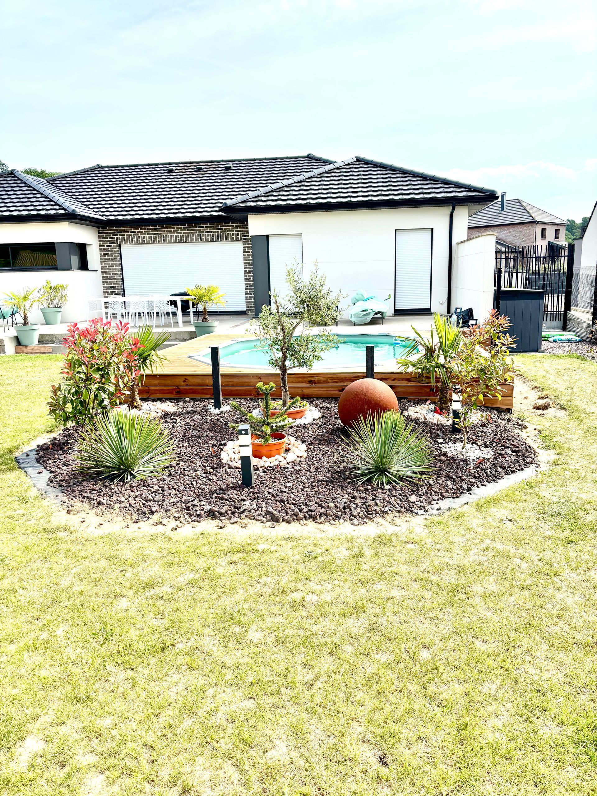 Un parterre de fleurs dans un jardin, un petit bassin et une maison blanche au toit de tuiles en arrière-plan.