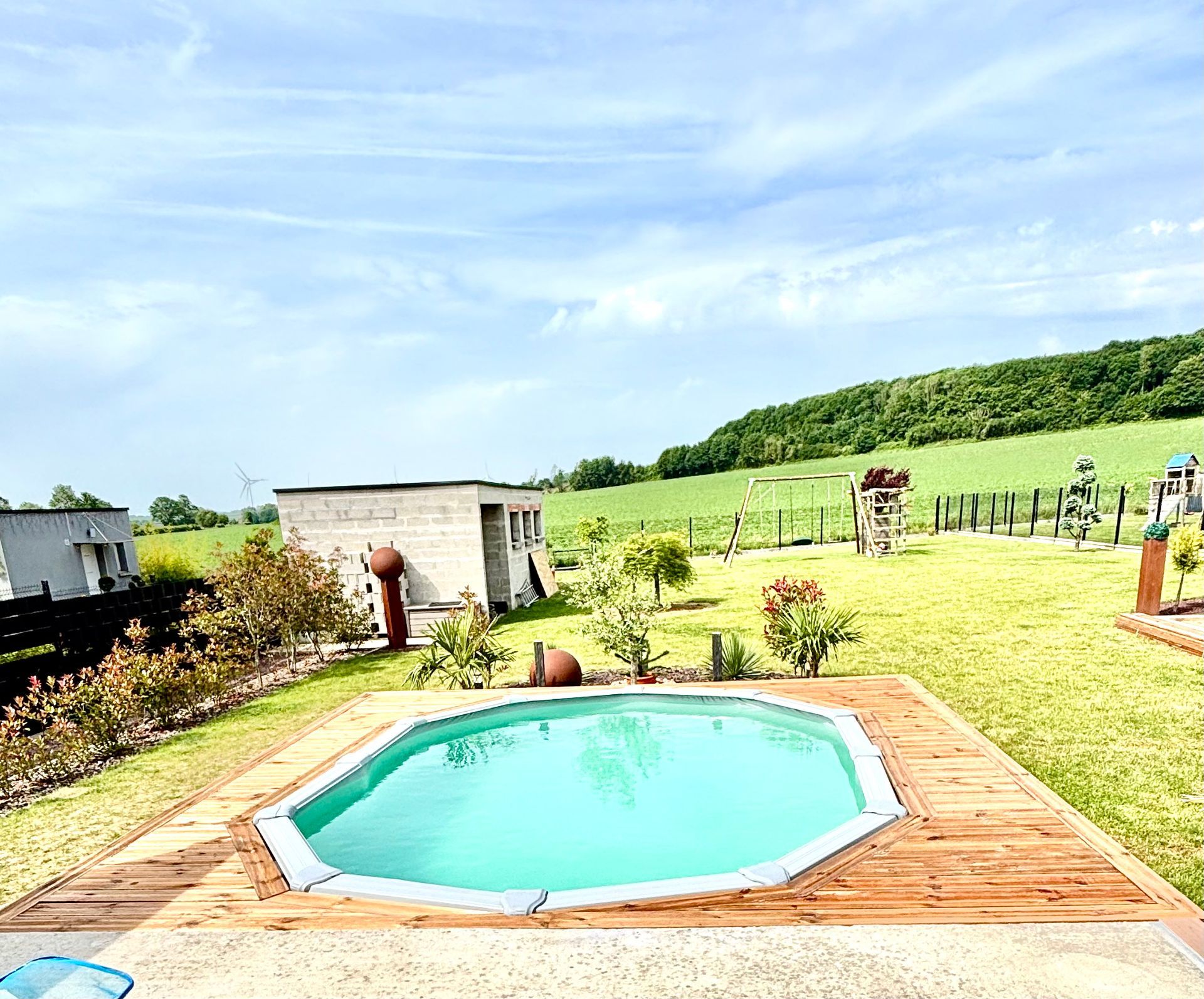 Terrasse arrière avec une petite piscine turquoise, une terrasse en bois, des parterres de fleurs et une colline herbeuse.