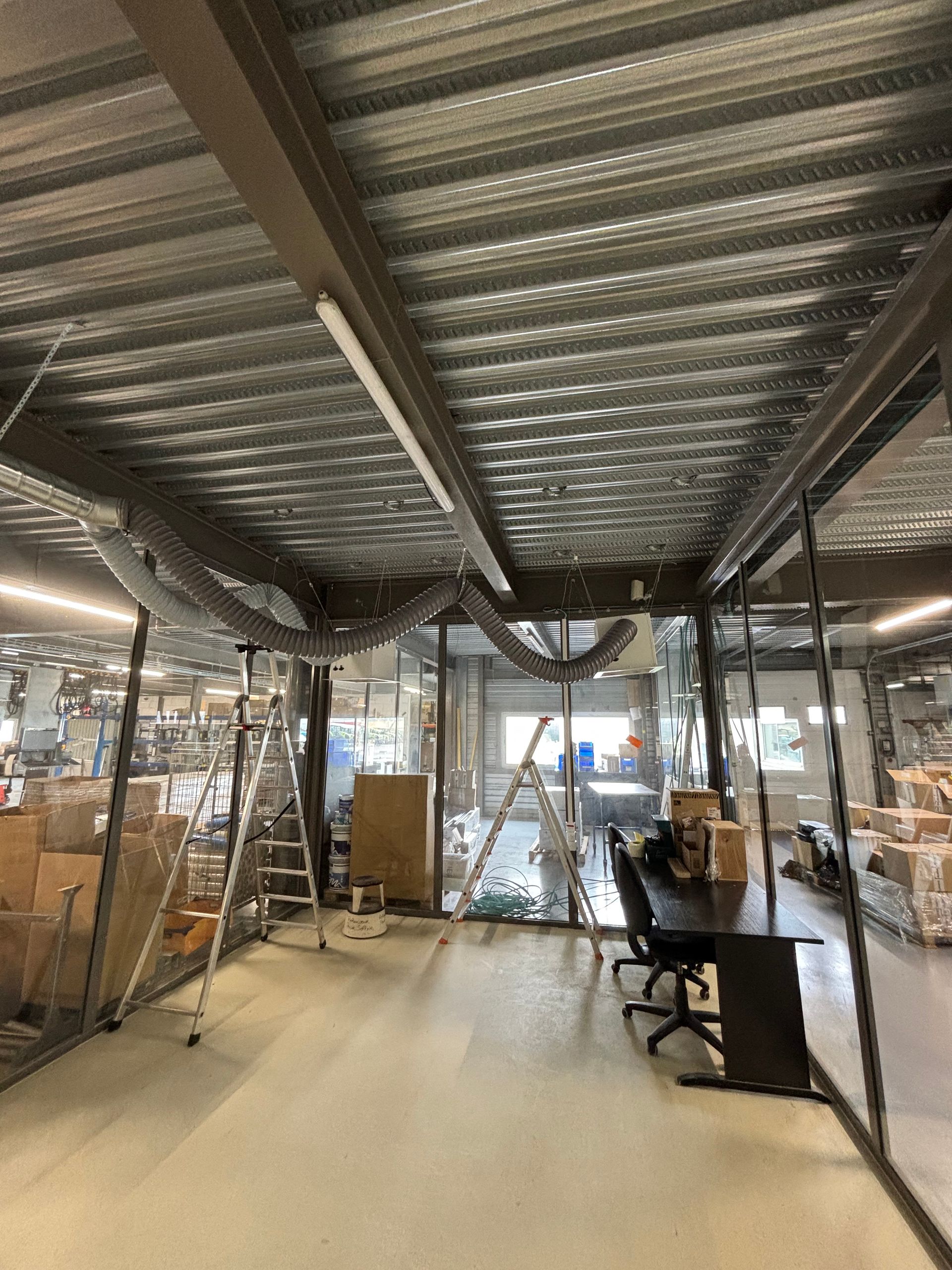 Intérieur d'un chantier de construction avec plafond apparent, des matériaux empilés et des postes de travail près de grandes fenêtres.