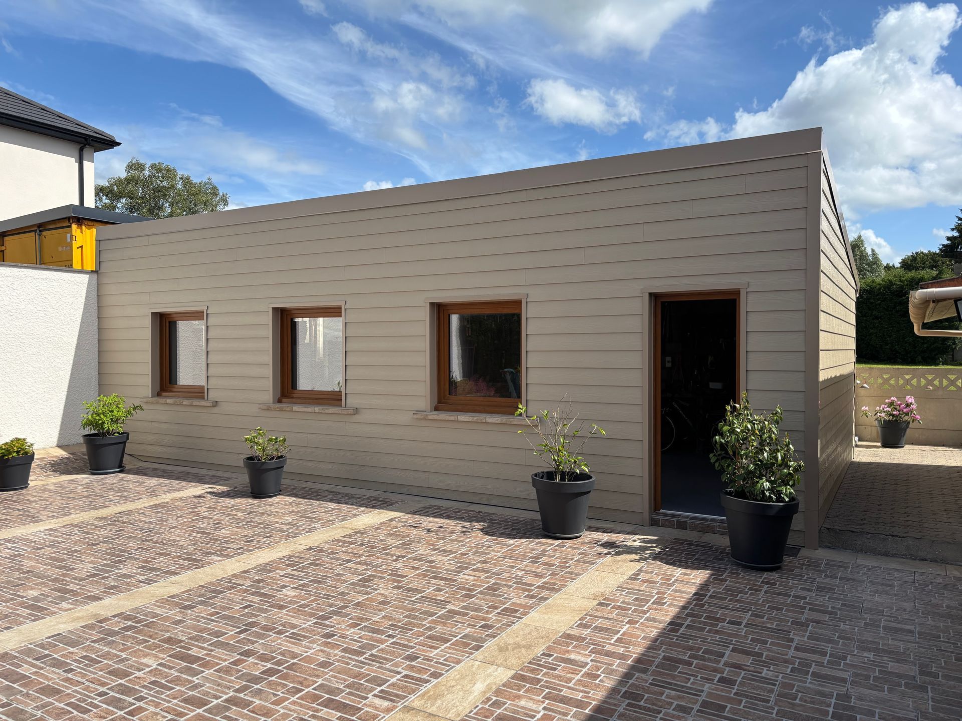 Bâtiment moderne beige avec trois fenêtres, porte noire, plantes en pot et cour en pierre à motifs.
