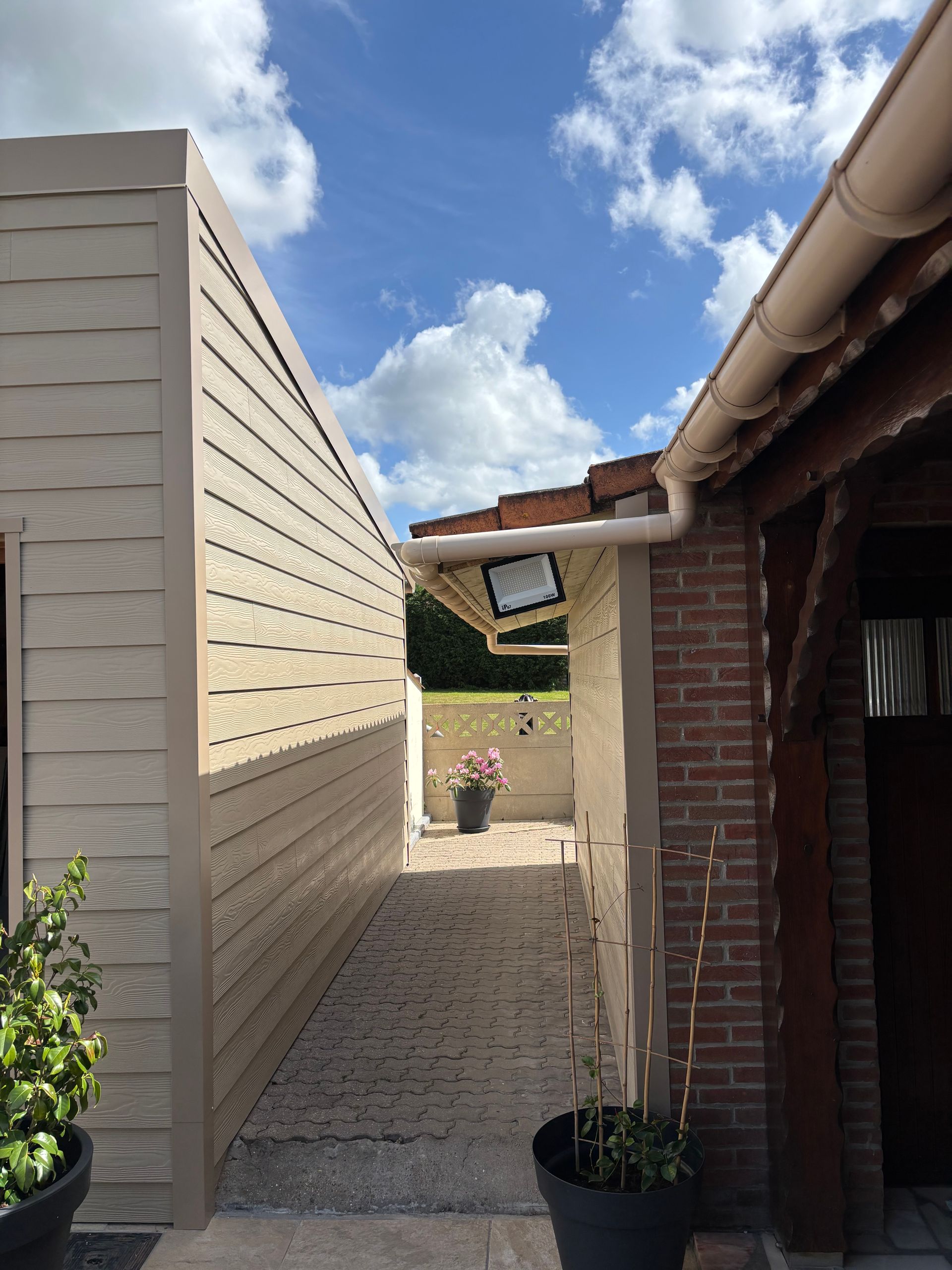 Un étroit chemin de gravier entre un abri beige et une maison en briques, menant à une cour herbeuse.