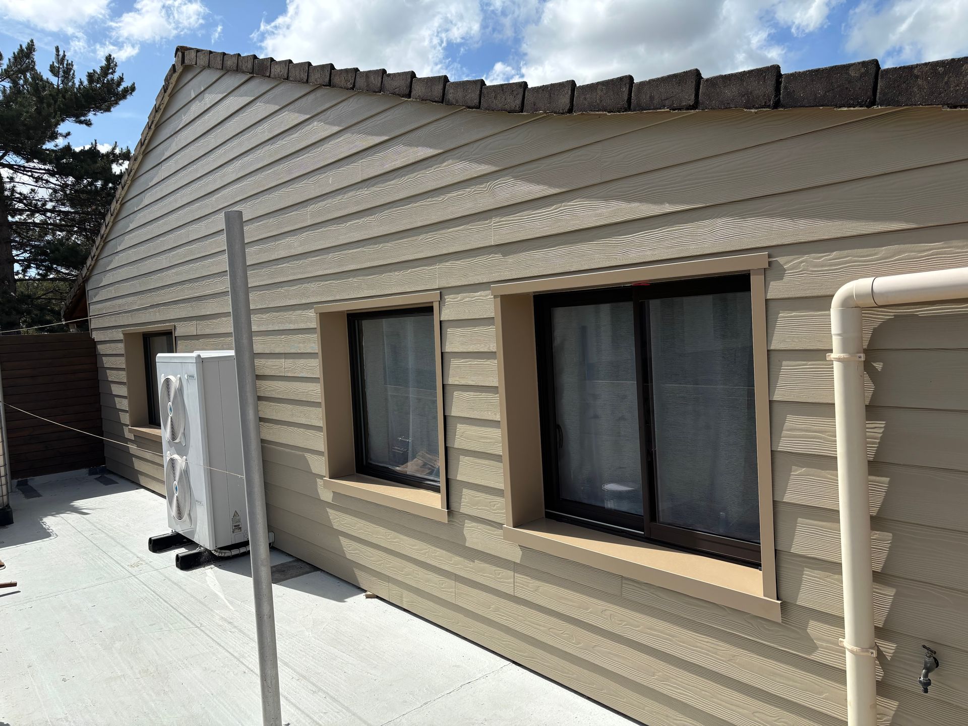 Extérieur d'une maison à bardage beige avec deux fenêtres et une unité de climatisation contre le mur.
