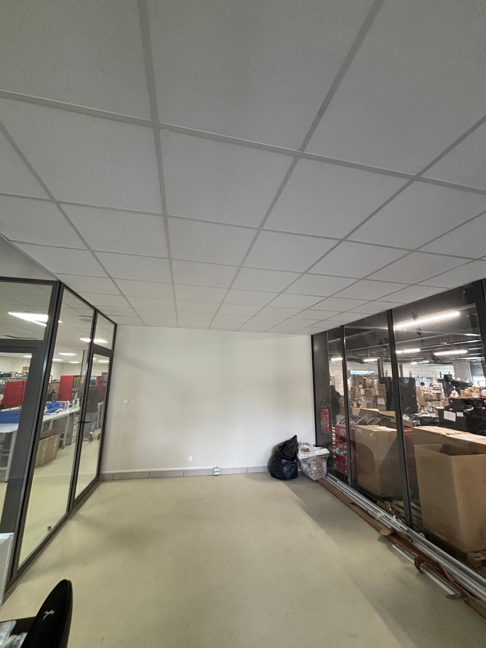 Couloir vide, semblable à un bureau, avec des murs en verre, des dalles de plafond blanches et des cartons empilés le long du côté droit.
