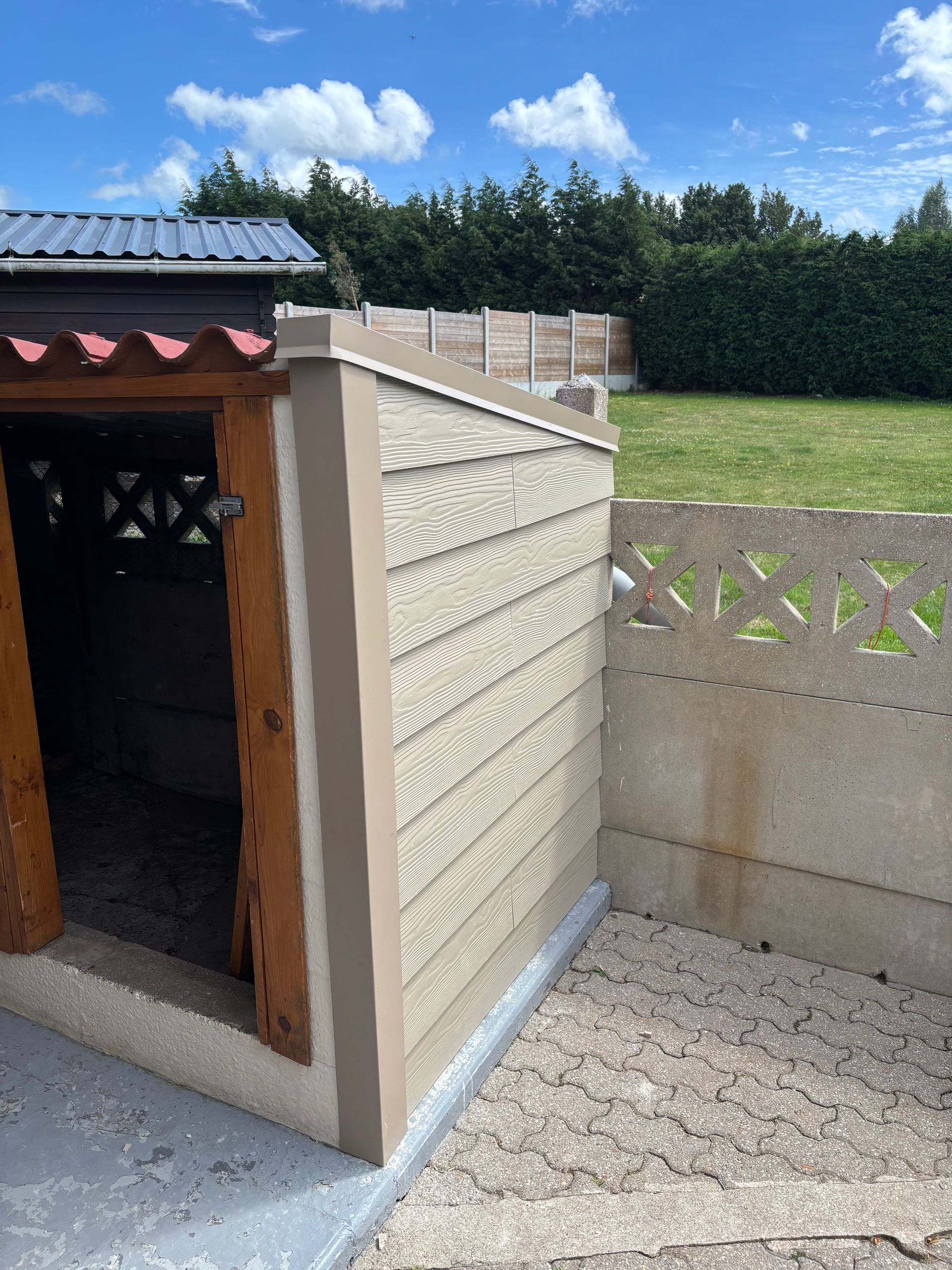 Niche extérieure pour chien avec un revêtement beige, une porte en bois et des murs en béton dans une cour ensoleillée.