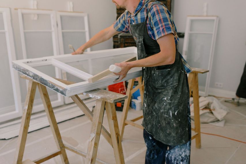 Une personne peint un cadre de fenêtre blanc dans un atelier, portant un tablier éclaboussé de peinture.