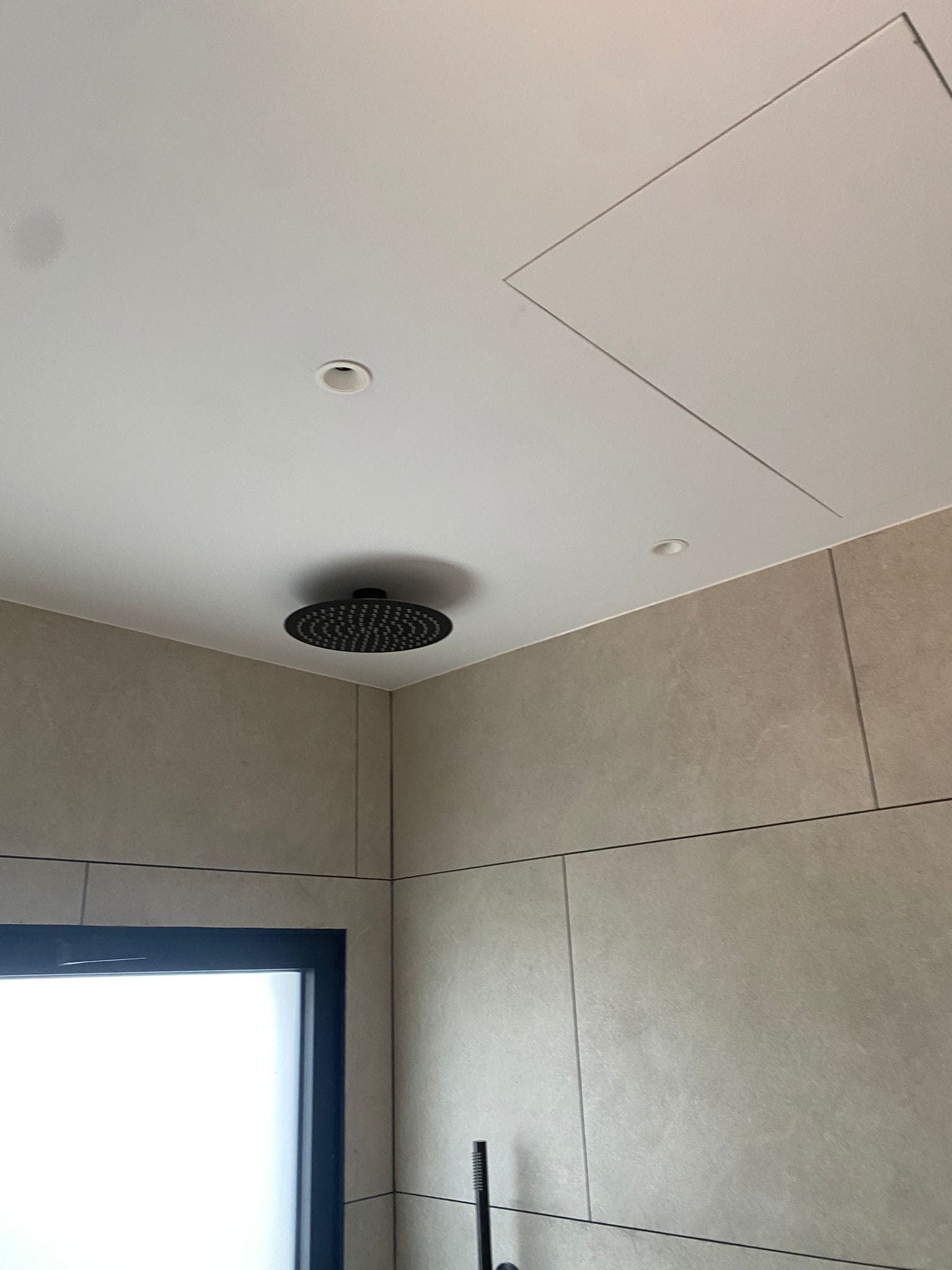 Ducha con cabezal circular negro sobre techo blanco. Se ven paredes beige y una ventana.