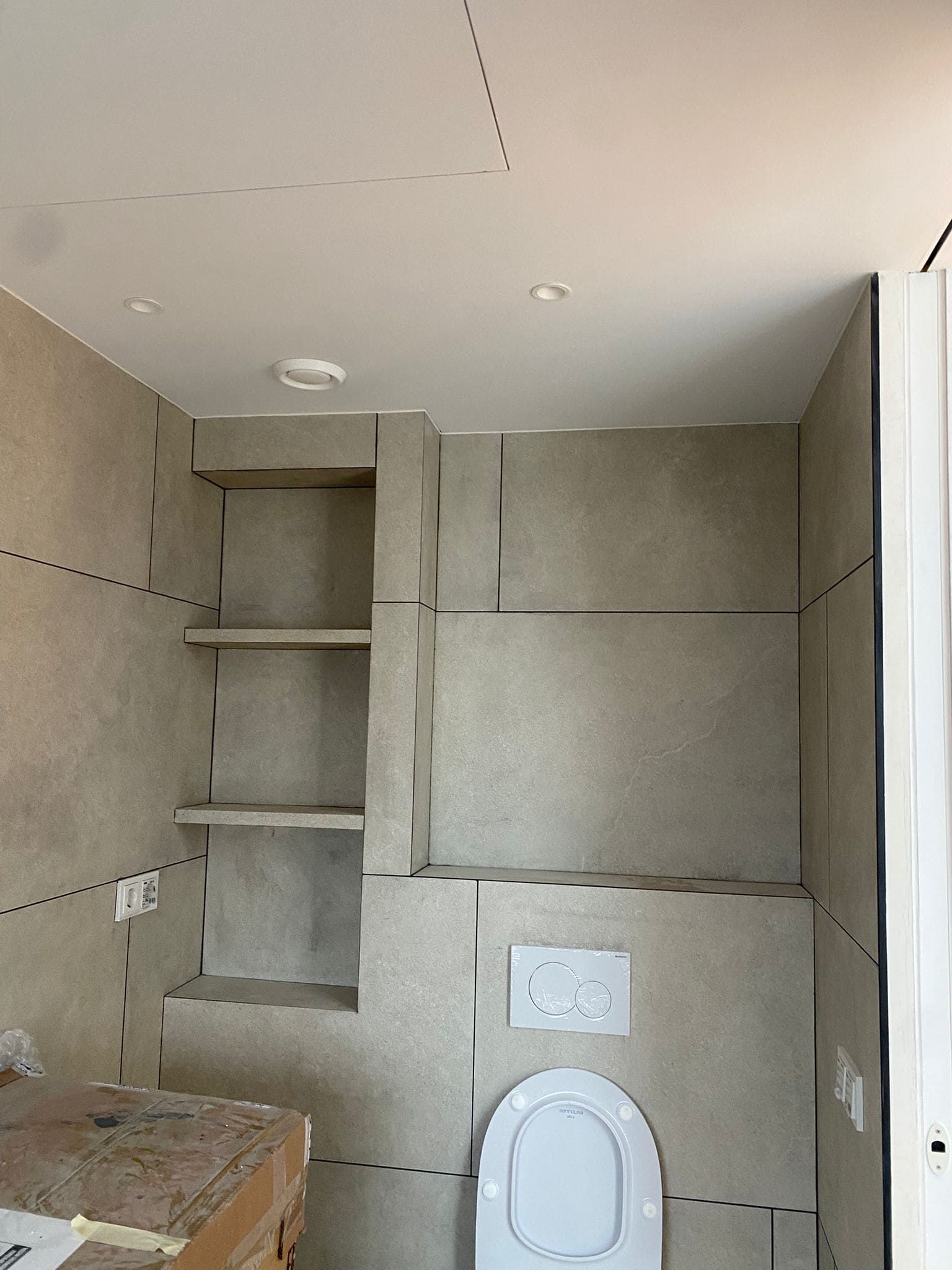 Baño en construcción, con inodoro, estantería empotrada y paneles de pared en color beige.