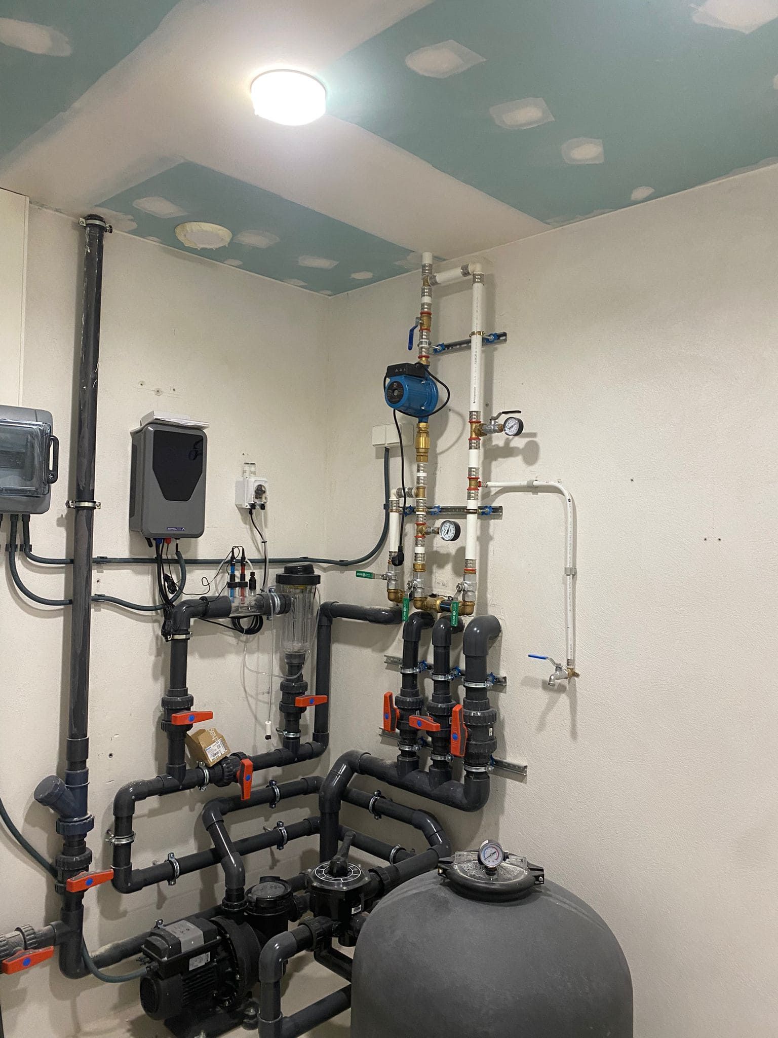 Sala de equipos de la piscina: tuberías, bombas y sistema de filtrado contra paredes blancas, con una luz de techo redonda.