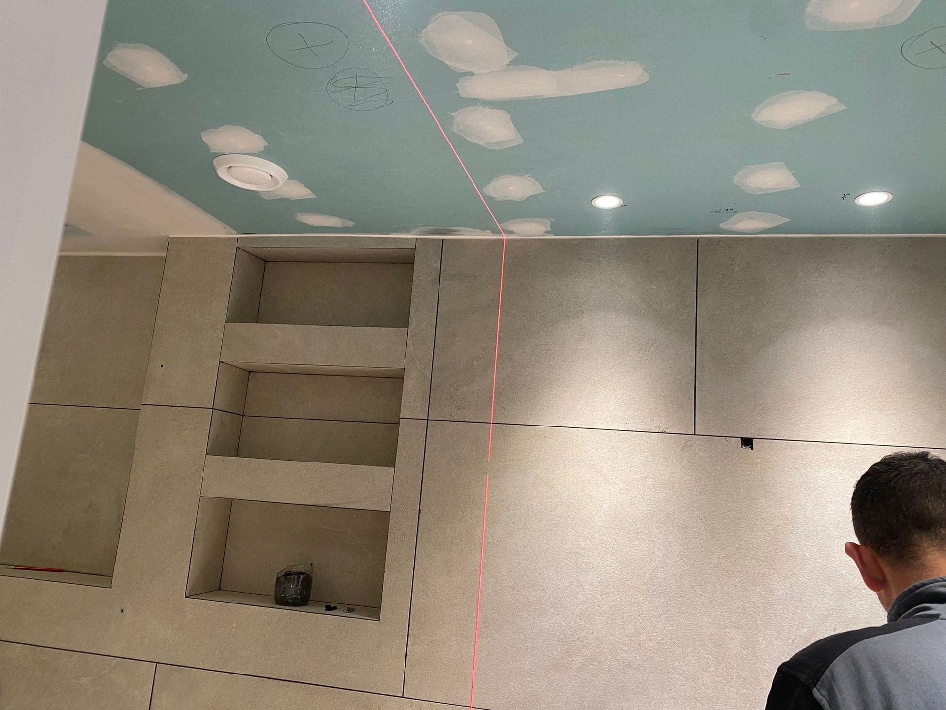 Habitación en construcción con estantes empotrados, paredes grises, techo azul claro, nivel láser, persona.