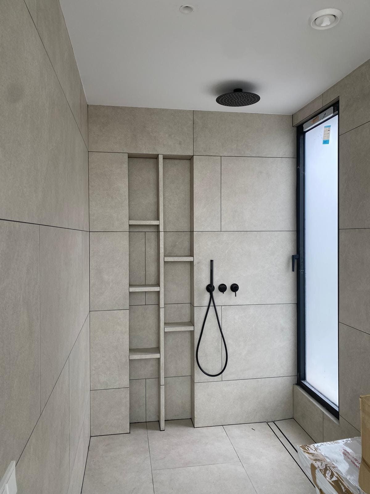 Ducha con azulejos grises, estante tipo nicho, accesorios negros y ventana esmerilada.