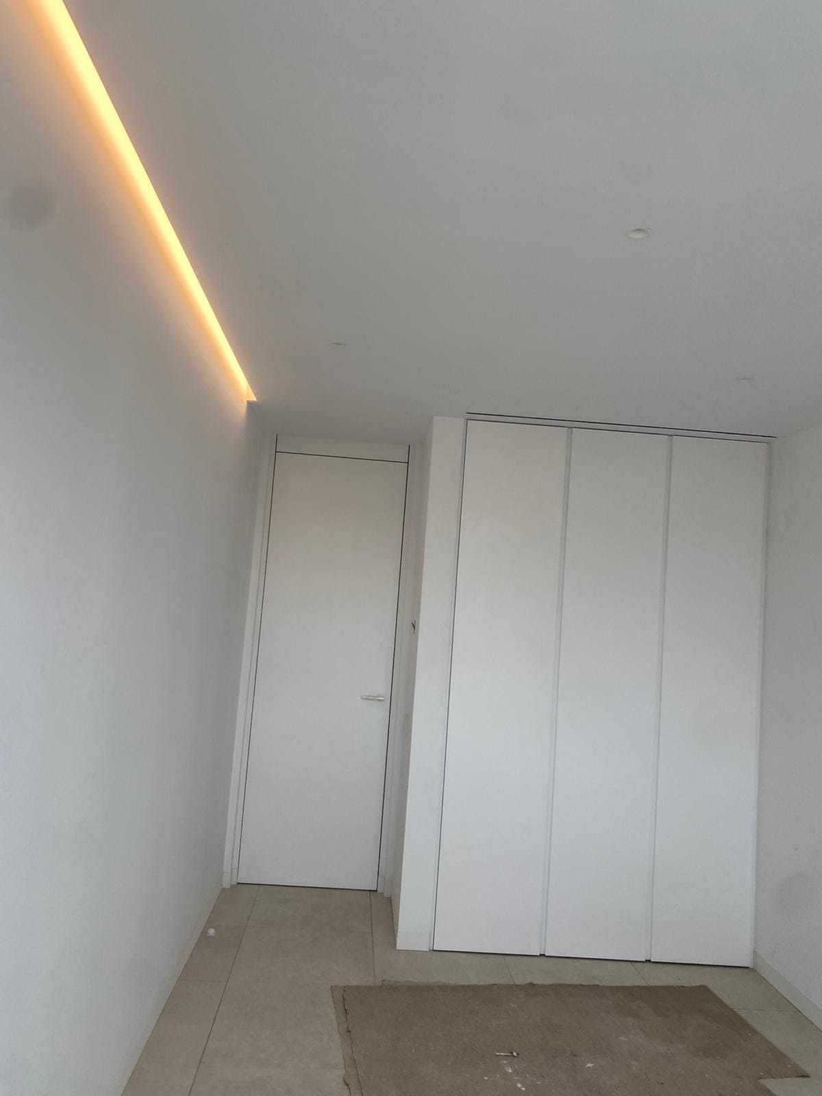 Habitación blanca con puerta, armario e iluminación empotrada. Hay una alfombra pequeña en el suelo.