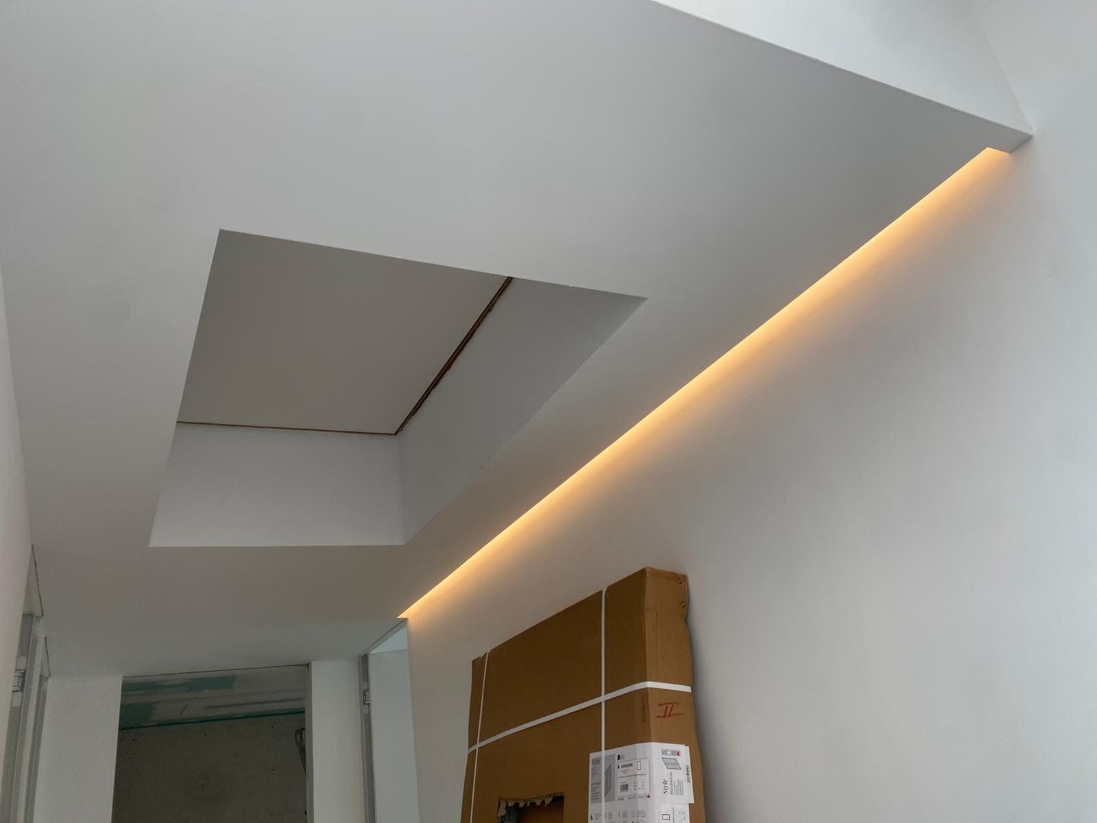 Techo blanco con sección empotrada y tira de iluminación LED en tonos cálidos; una caja de cartón reposa en la pared.