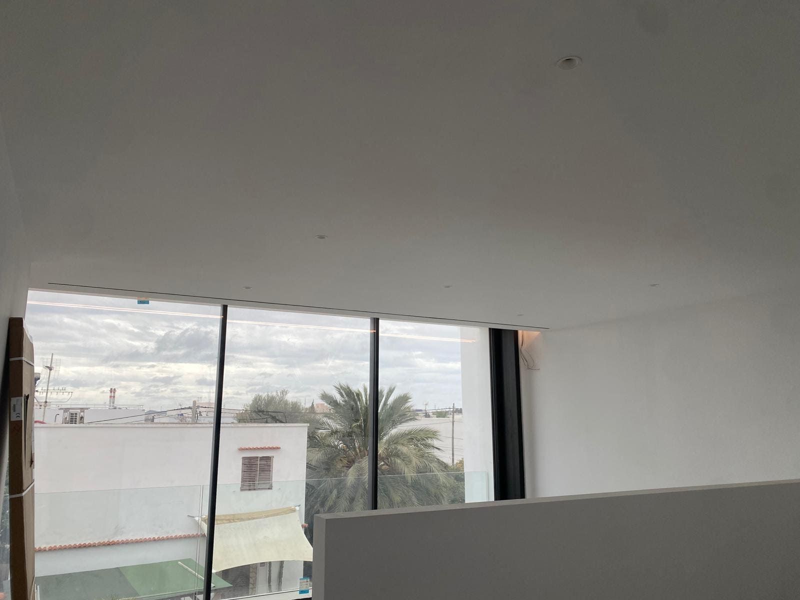 Vista interior de una ventana con vistas a edificios urbanos y palmeras. Cielo nublado y brillante.