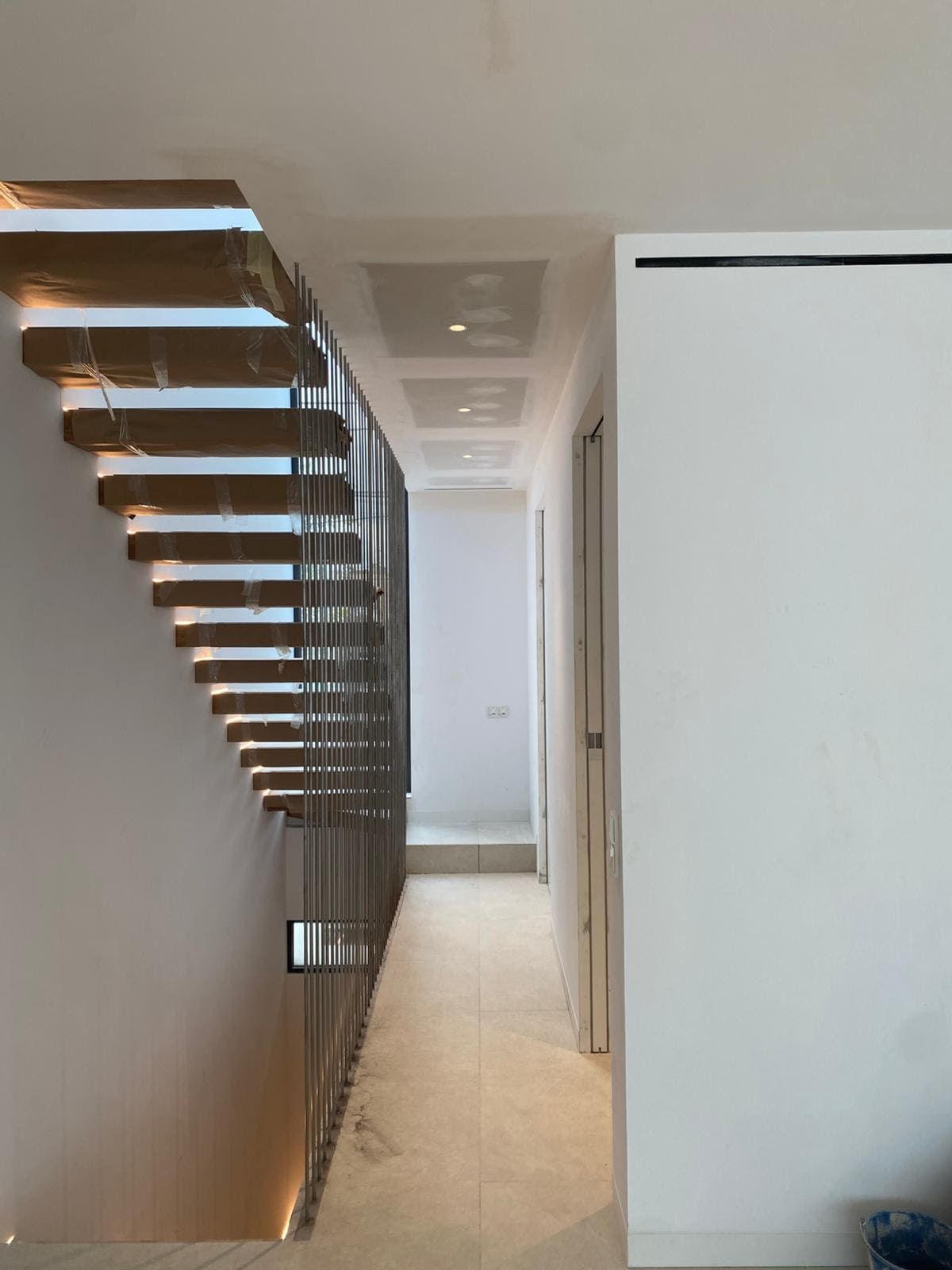 Escalera de madera con barandilla metálica junto a un pasillo con paredes blancas y una puerta.