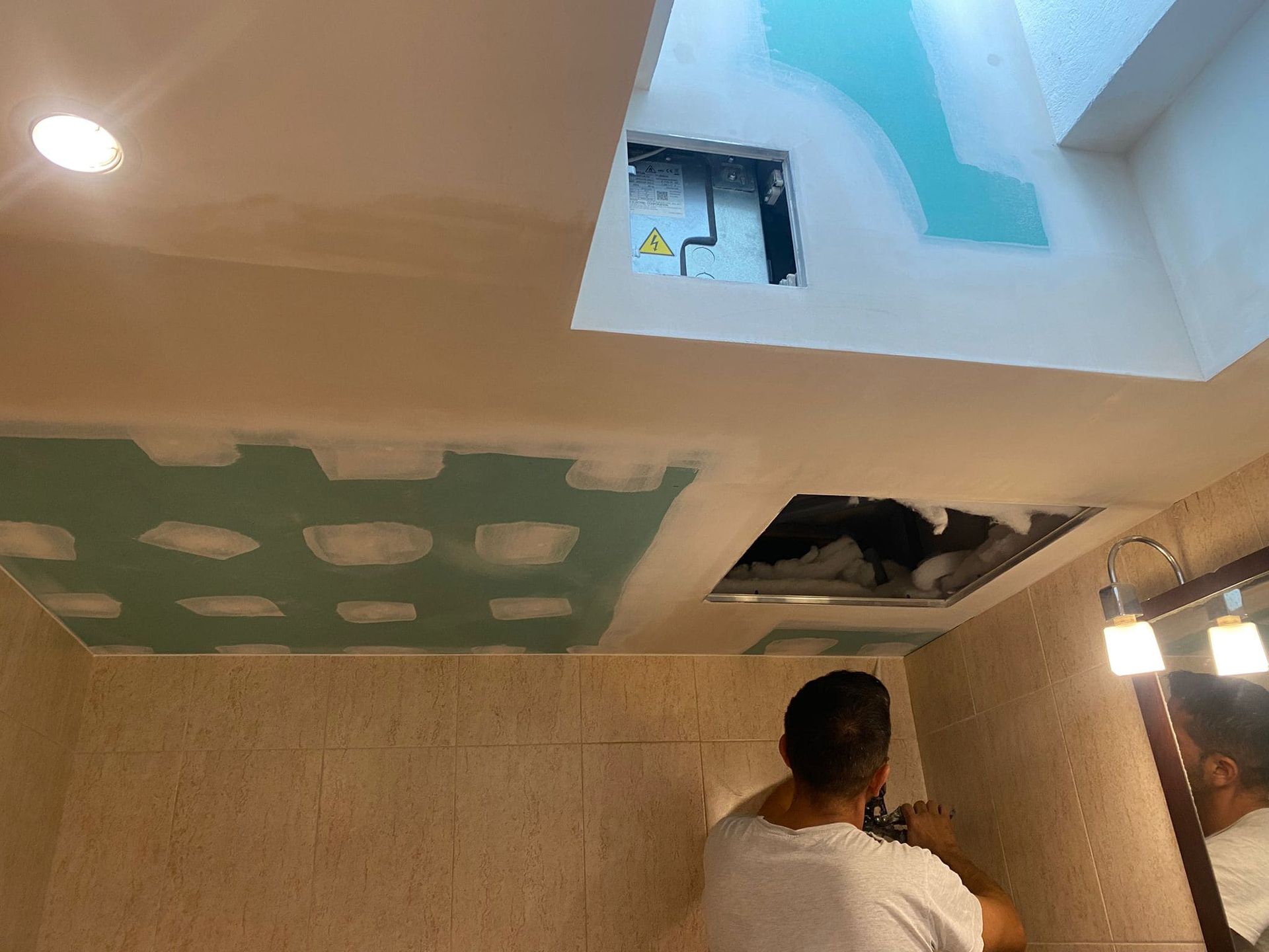 Hombre trabajando en el techo del baño, instalando paneles de yeso. Se ven el panel del techo, las lámparas y el espejo.