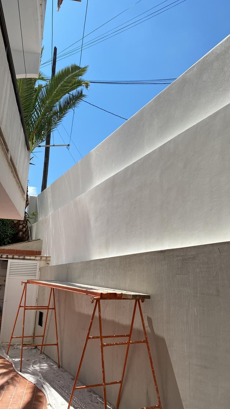 Una pared alta y blanca con una franja de luz, una escalera, cables eléctricos y una palmera contra un cielo azul brillante.