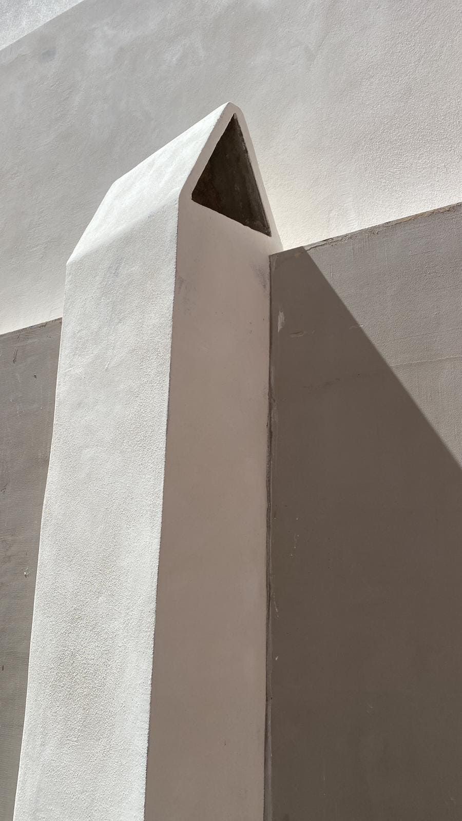 Chimenea angular blanca con abertura triangular, adosada a una pared texturizada; iluminada por la luz del sol, proyectando sombras.